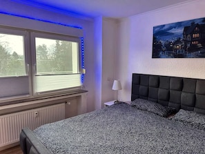 A bedroom with a large bed, a window with blinds, a radiator, and a painting on the wall.