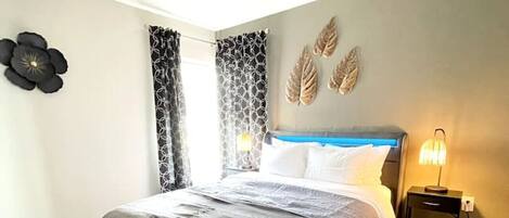 A bedroom with a bed, curtains, a wall clock, and a nightstand with a lamp.