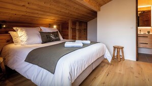 A bedroom with a wooden ceiling, a bed with white bedding, a grey throw, and a pillow with text. There is a wooden stool and a wooden door leading to a bathroom. - Le Primpiolet - Penthouse for 9 with Mont Blanc view - OVO Network (Combloux)