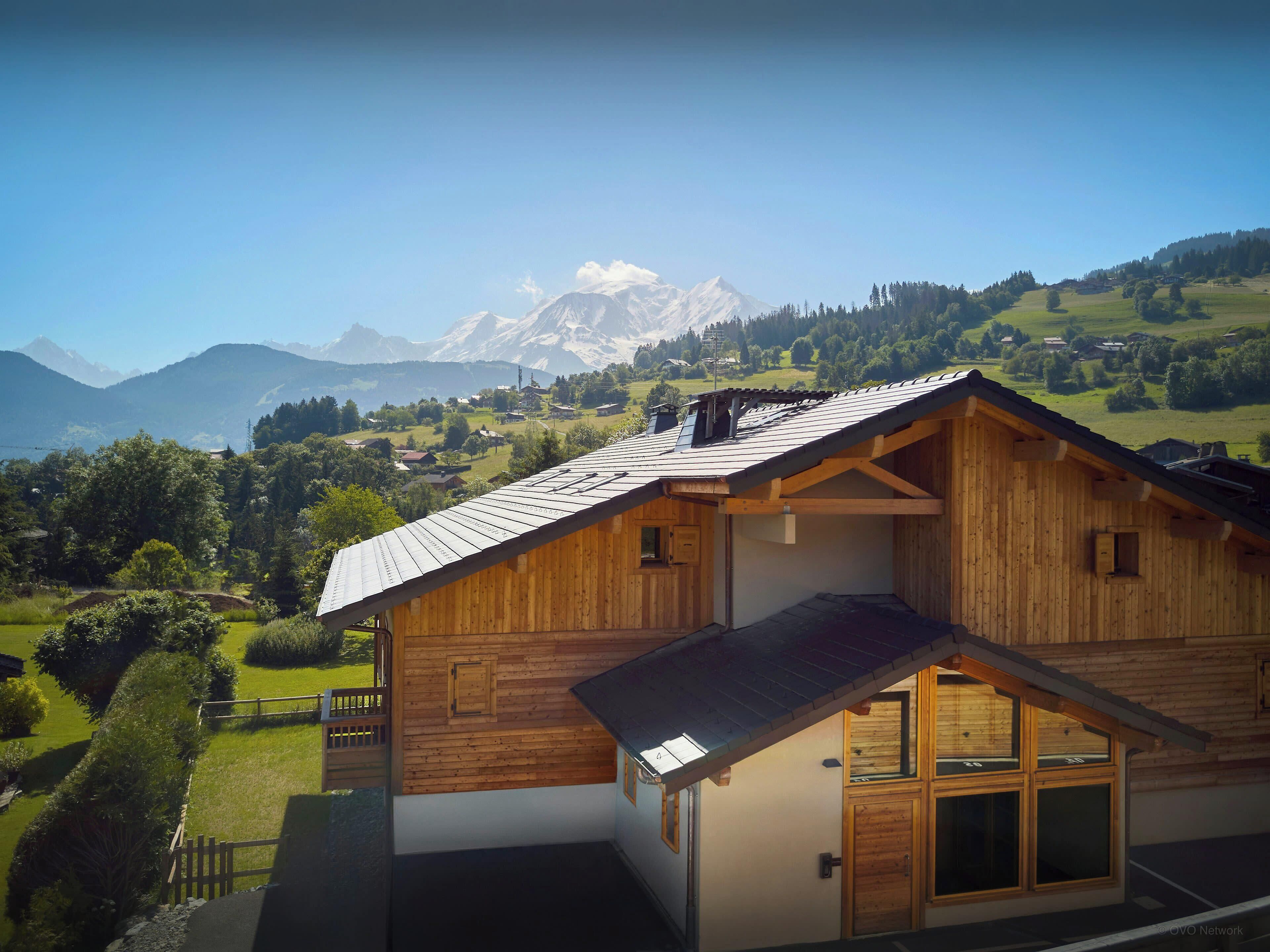 A wooden chalet with a sloped roof, large windows, and a view of a mountain range.