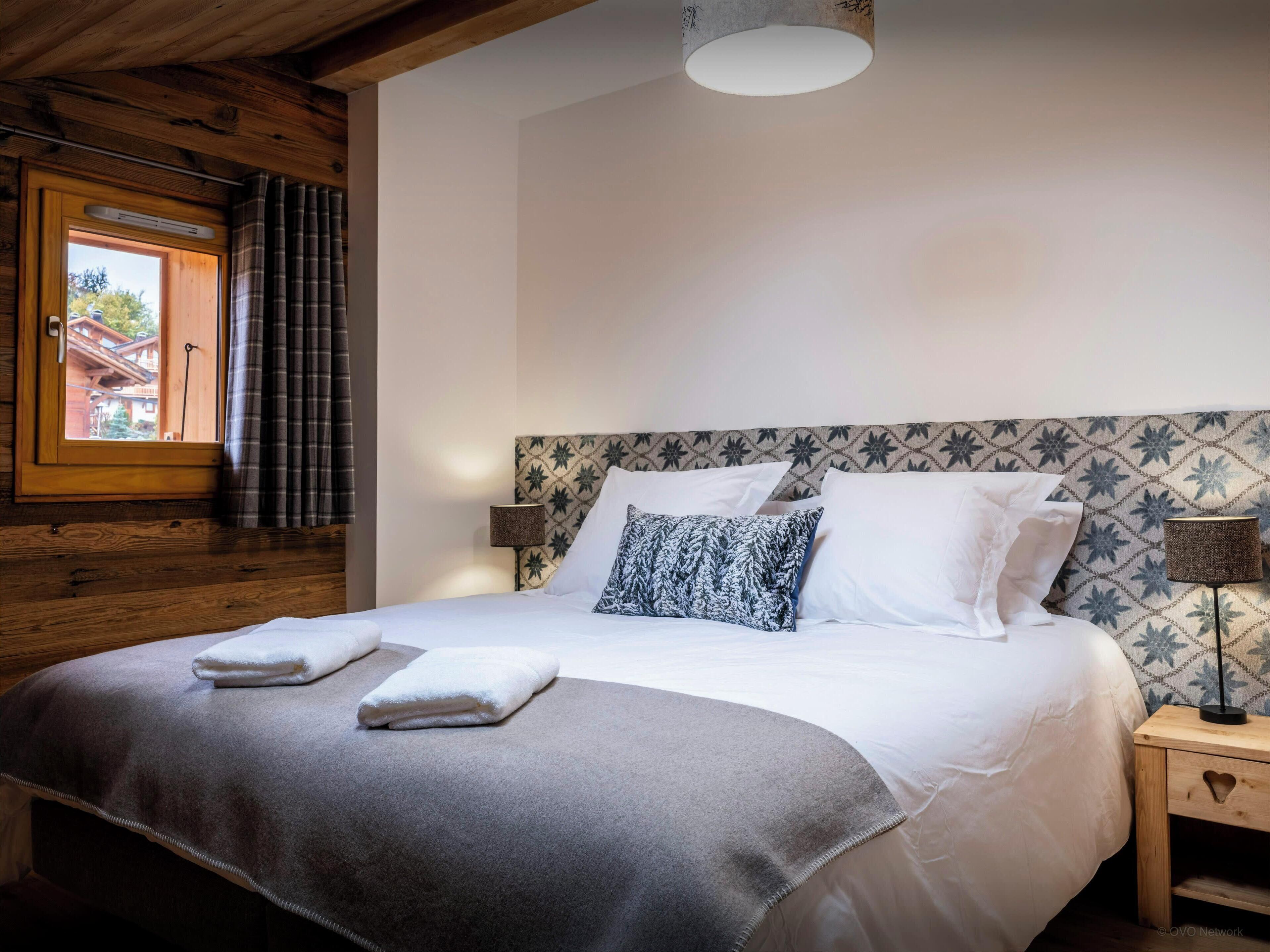 A neatly made bed with white and patterned pillows, a grey blanket, and two white towels. There is a window with curtains, a wall-mounted lamp, and a patterned headboard.