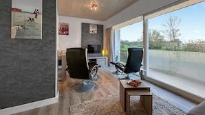 Un salon moderne avec une grande fenêtre, deux fauteuils noirs, une petite table en bois et un téléviseur posé sur un meuble.