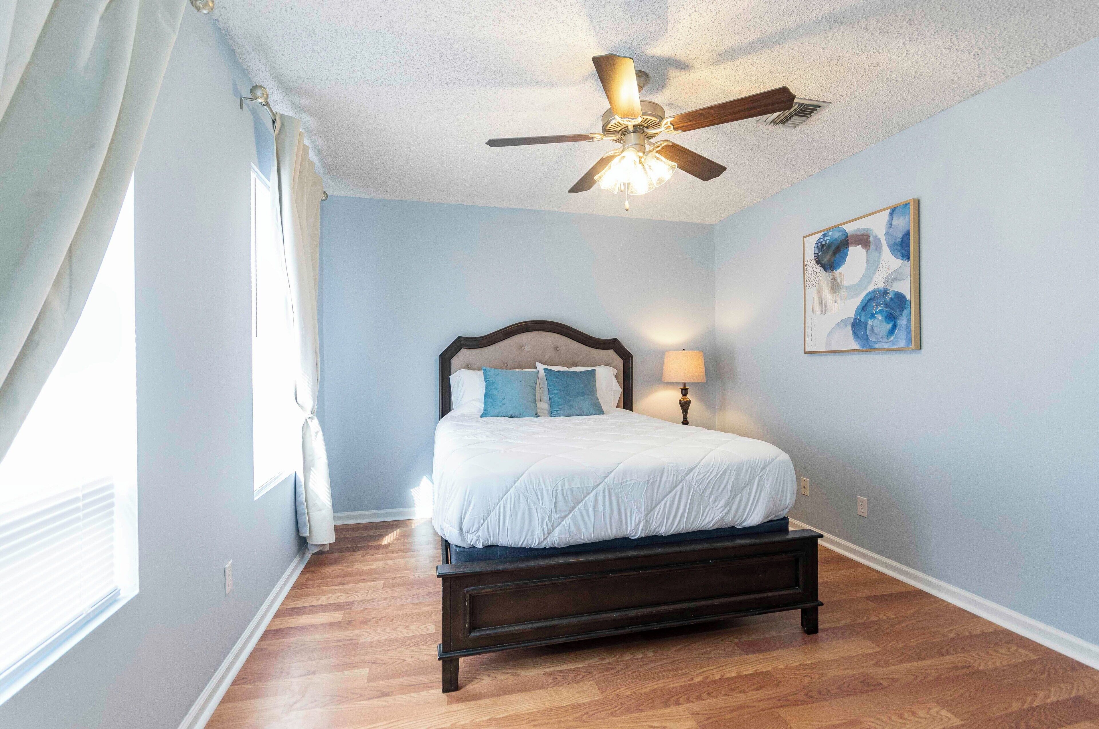 A bedroom with a bed, a ceiling fan, a painting, and a window with curtains.