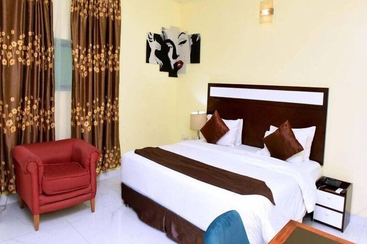 A hotel room with a bed, a red armchair, a nightstand, and curtains with a floral pattern.