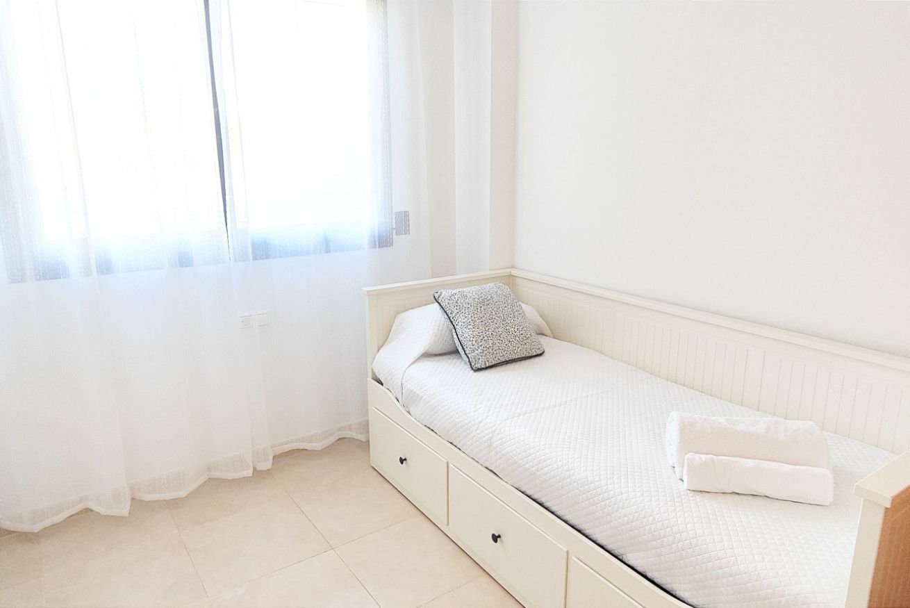 A single bed with a white mattress and pillows, a bedside table, and a window with sheer curtains.