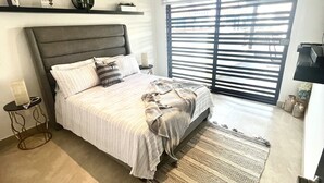 Quarto com cama, mesa de cabeceira, mesinha, janela com persianas e prateleira com objetos decorativos.