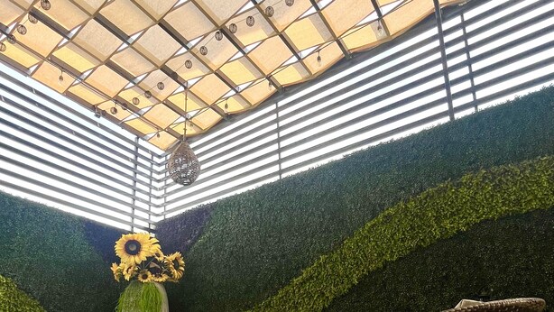 Uma área de estar coberta ao ar livre com móveis de vime, um vaso grande com girassóis e um painel de parede verde.