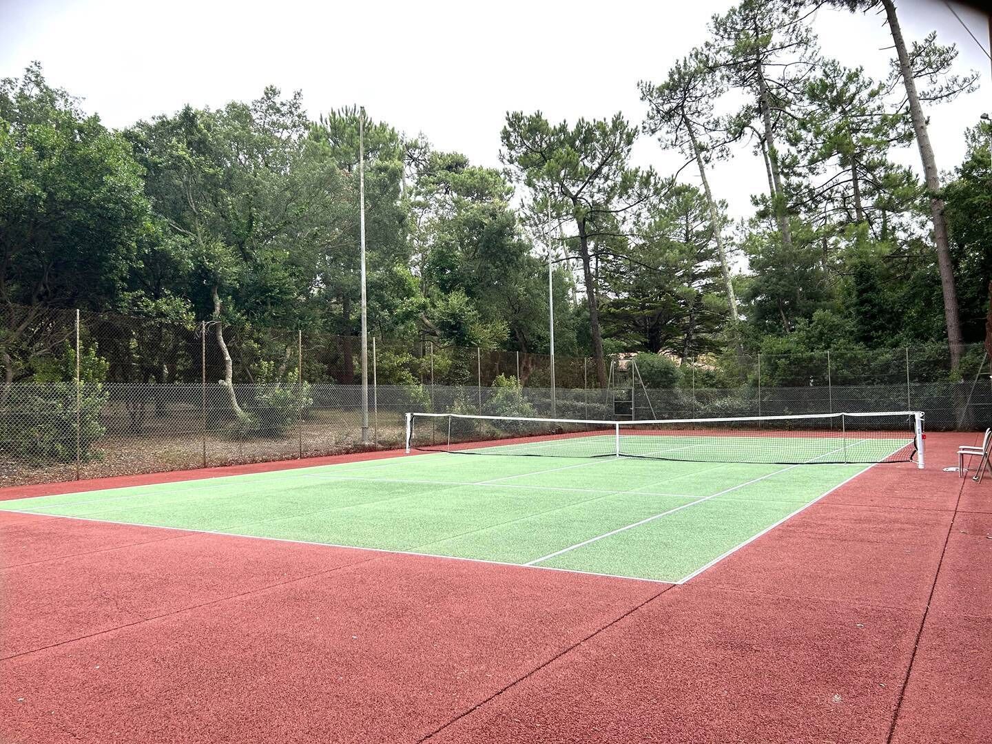 An outdoor tennis court with a red surface, surrounded by a fence and trees.