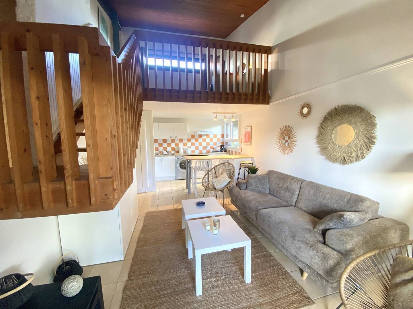 A modern living room with a grey sofa, white coffee table, and wicker chairs. A kitchen with a checkered backsplash is visible in the background.