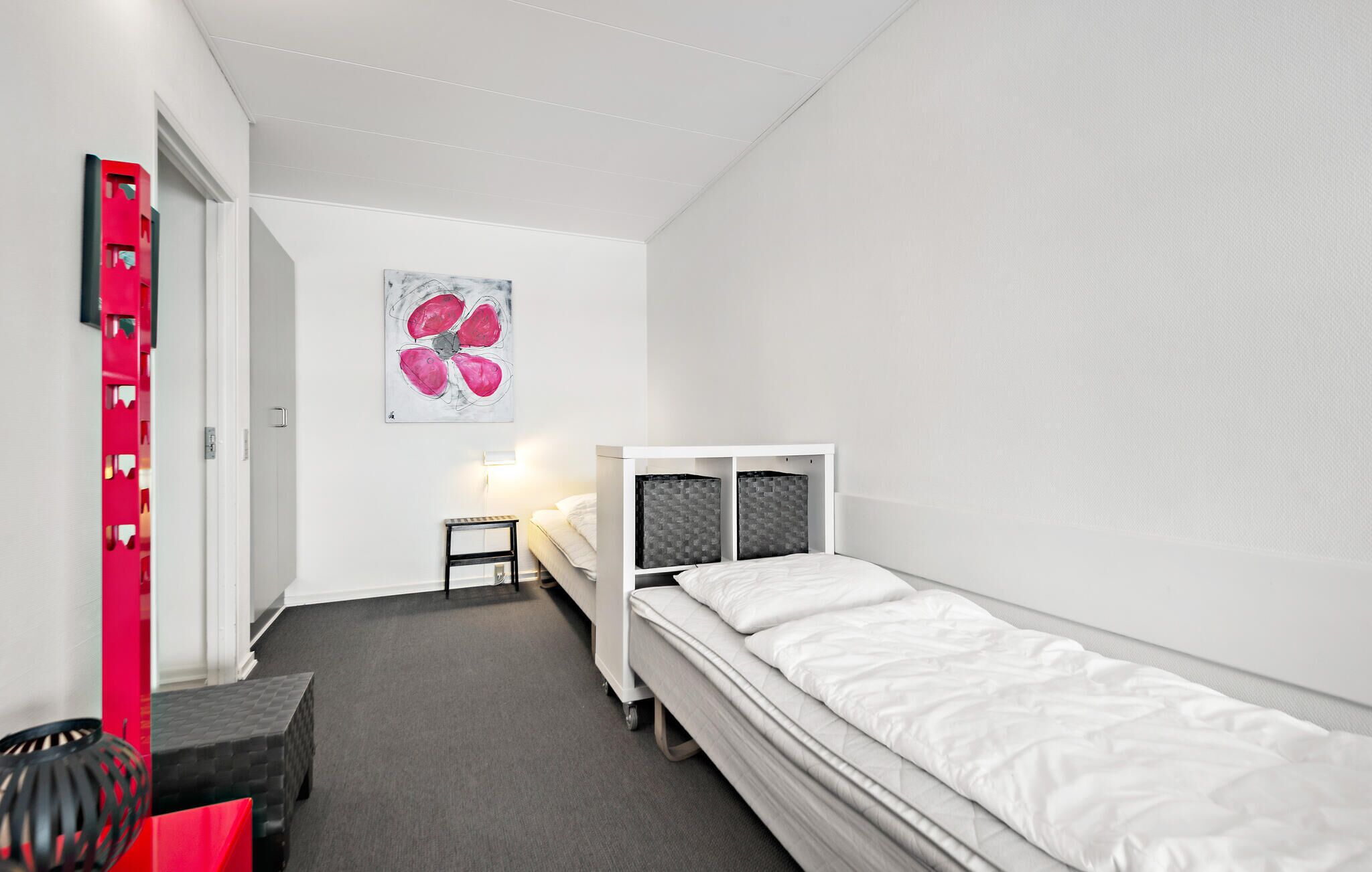 Quarto pequeno e moderno com uma cama de solteiro, uma escrivaninha e uma cadeira. Há uma pintura na parede e um objeto vermelho no lado esquerdo.