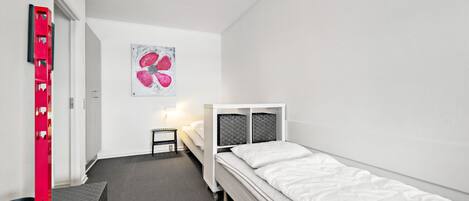 Quarto pequeno e moderno com uma cama de solteiro, uma escrivaninha e uma cadeira. Há uma pintura na parede e um objeto vermelho no lado esquerdo.