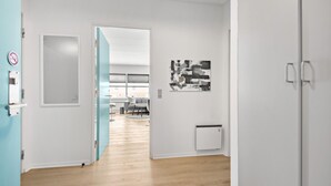 Un corridoio moderno e luminoso con pavimento in legno che conduce a un soggiorno con divano e tavolino. Si nota una porta con interruttore della luce e un'opera d'arte appesa al muro.
