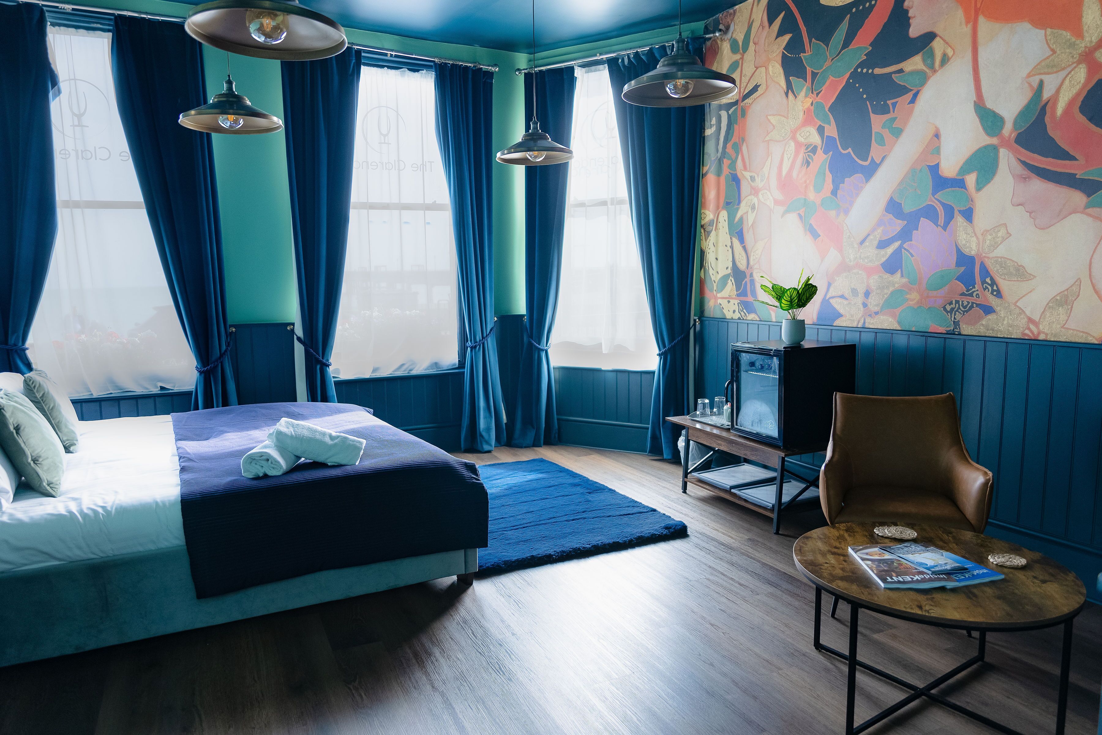 Quarto de hotel moderno com uma cama grande, uma mesa pequena com revistas, uma cadeira e uma janela ampla com cortinas.