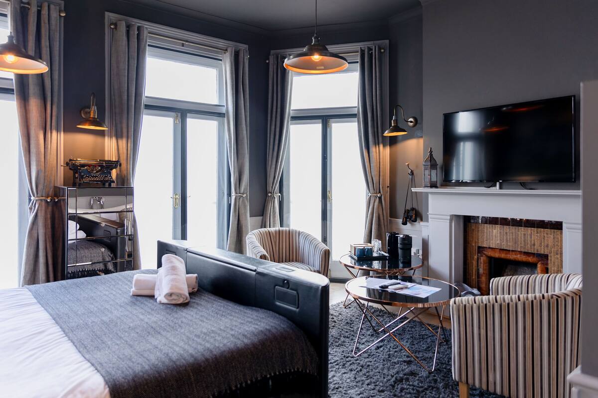A modern hotel room with a large bed, a TV, a fireplace, and a seating area.