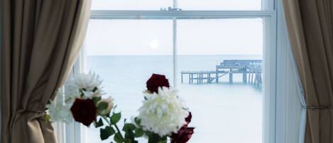 A room with a round white table, a vase with flowers, and a view of the sea through a large window.