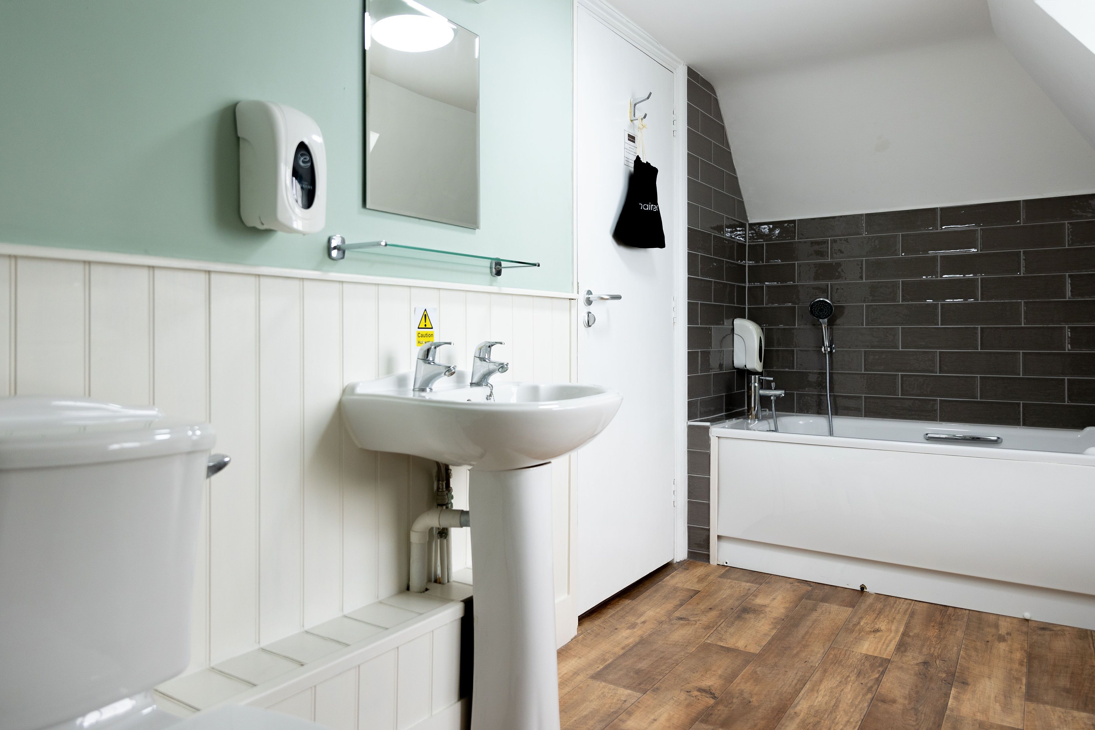 A bathroom with a white sink, mirror, and toilet, a black shower area, and wooden flooring.