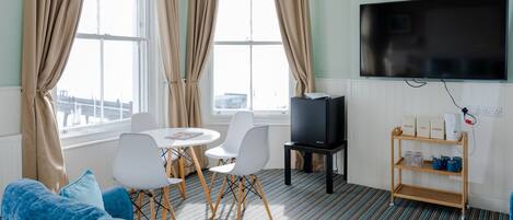 A modern hotel room with a large TV, a dining table with chairs, a sofa, and a coffee station.