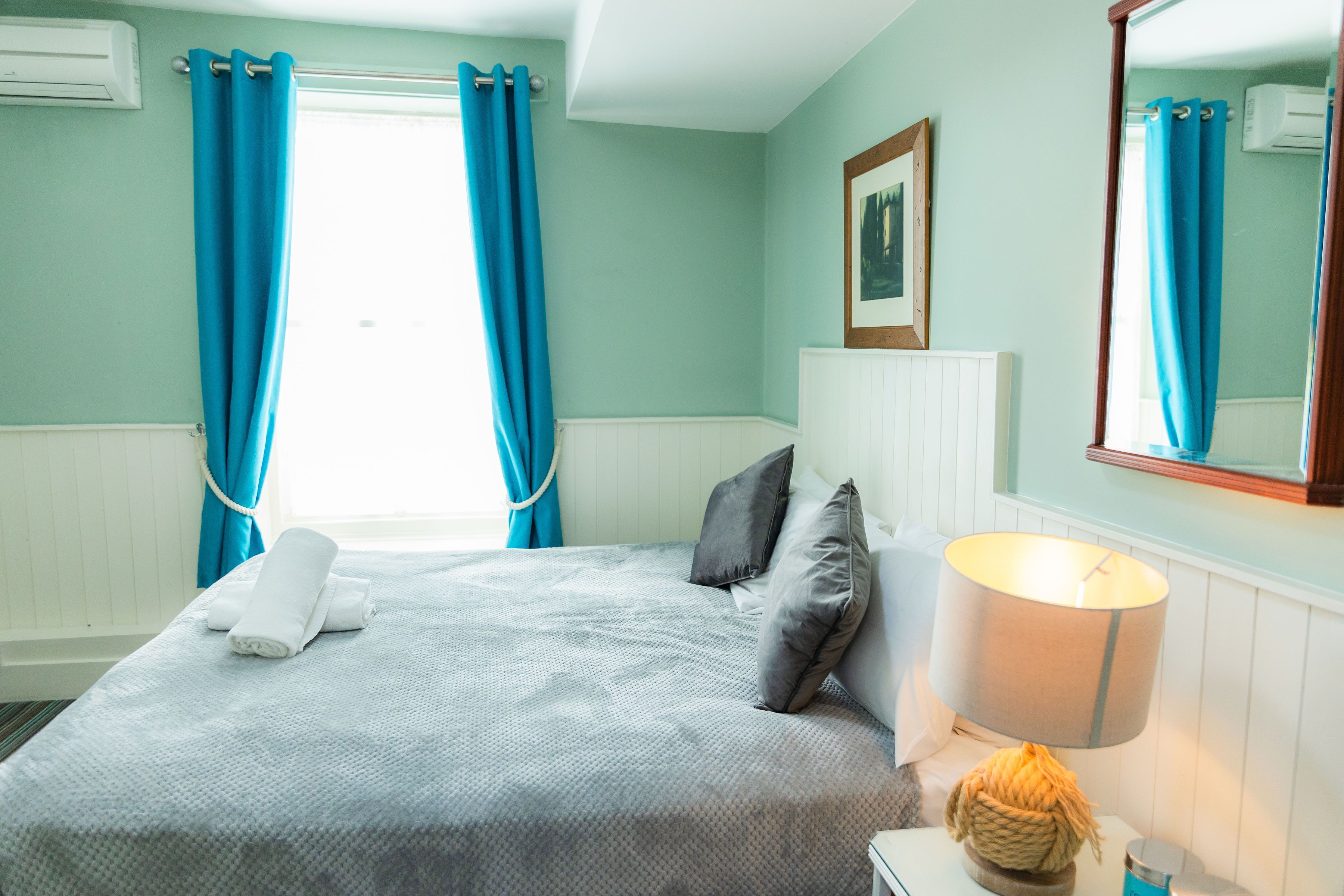 A neatly made bed with a white comforter and pillows, a bedside lamp, and a window with blue curtains.
