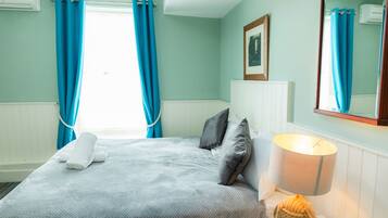 A neatly made bed with a white comforter and pillows, a bedside lamp, and a window with blue curtains.