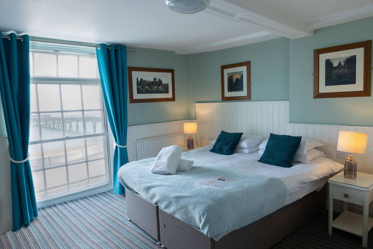 A neatly made bed with a white comforter and teal pillows, a bedside table with a lamp, and a window with curtains.