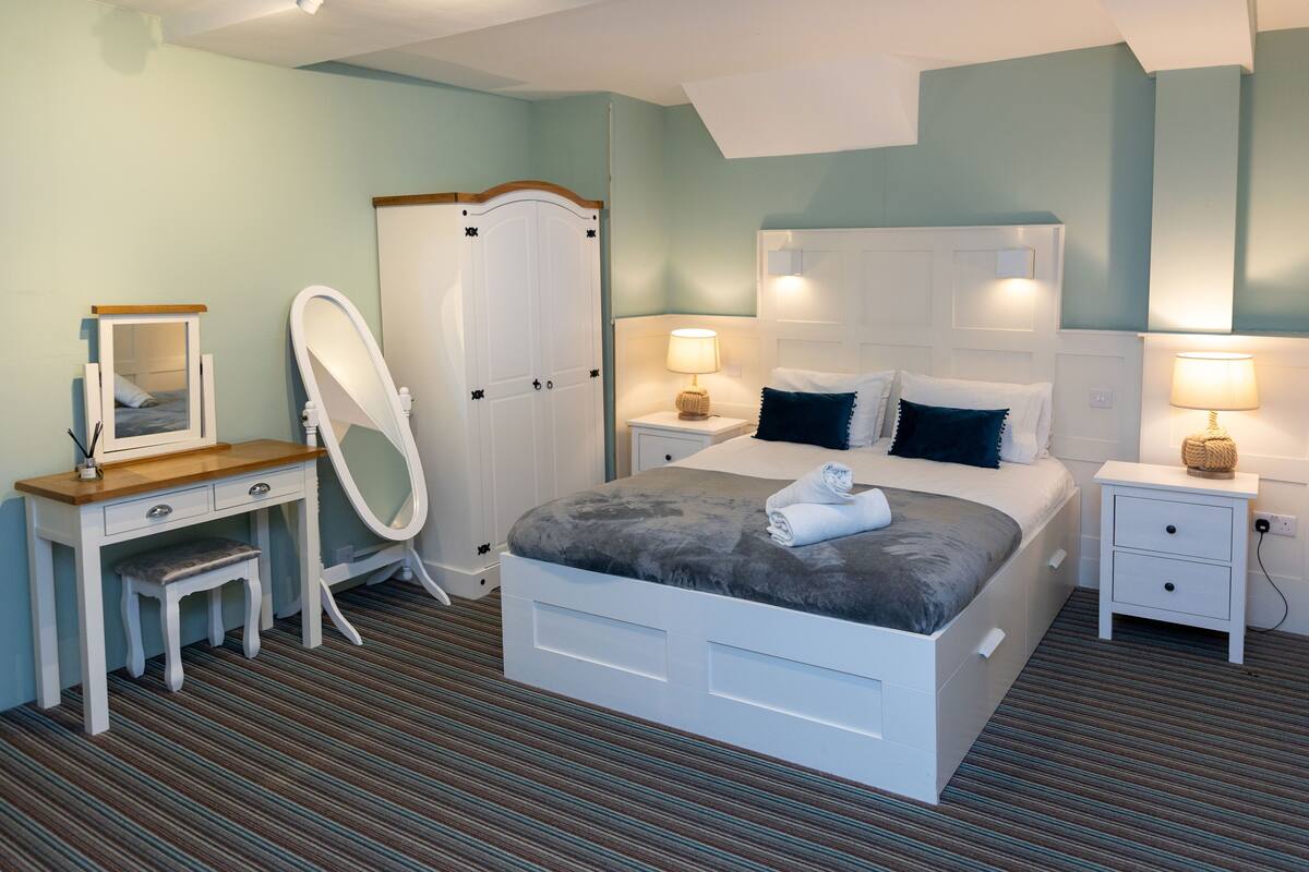 A neatly arranged bedroom with a bed, bedside tables, a mirror, a wardrobe, and a small stool.