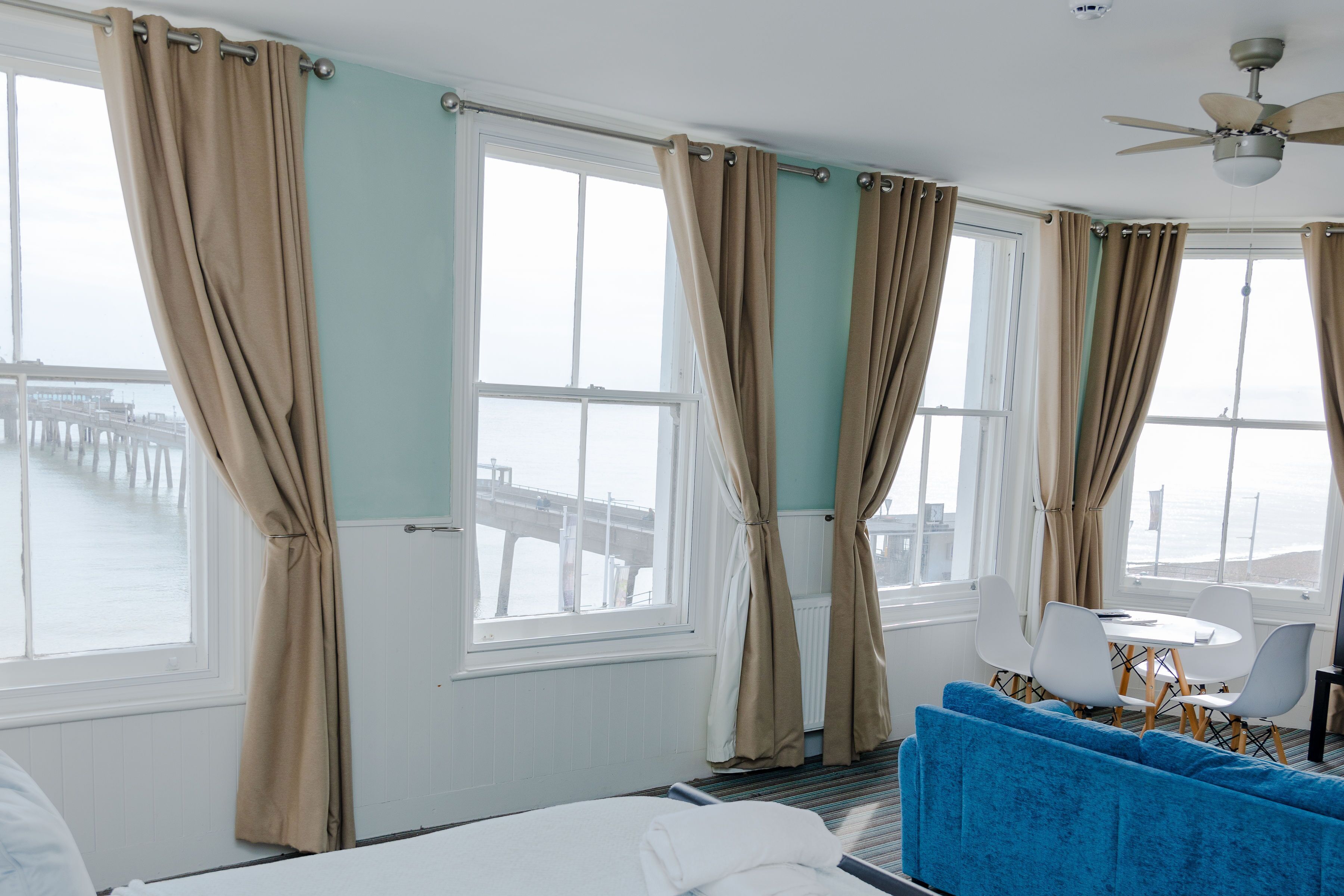 Ein Hotelzimmer mit einem Bett, einem blauen Sofa, einem Esstisch mit Stühlen und großen, mit Vorhängen versehenen Fenstern.