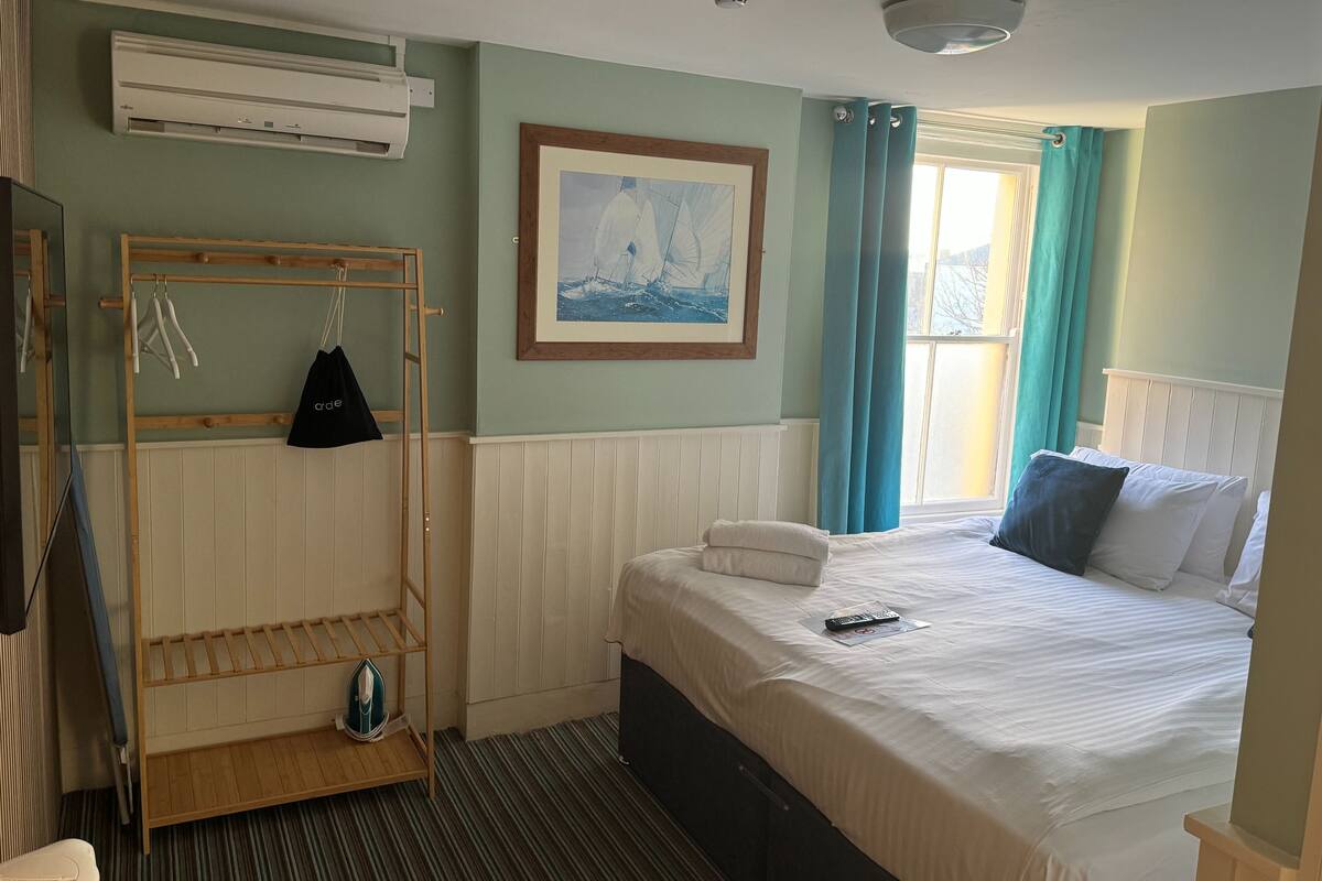 A hotel room with a bed, a hanging clothes rack, a framed picture, and a window with curtains.