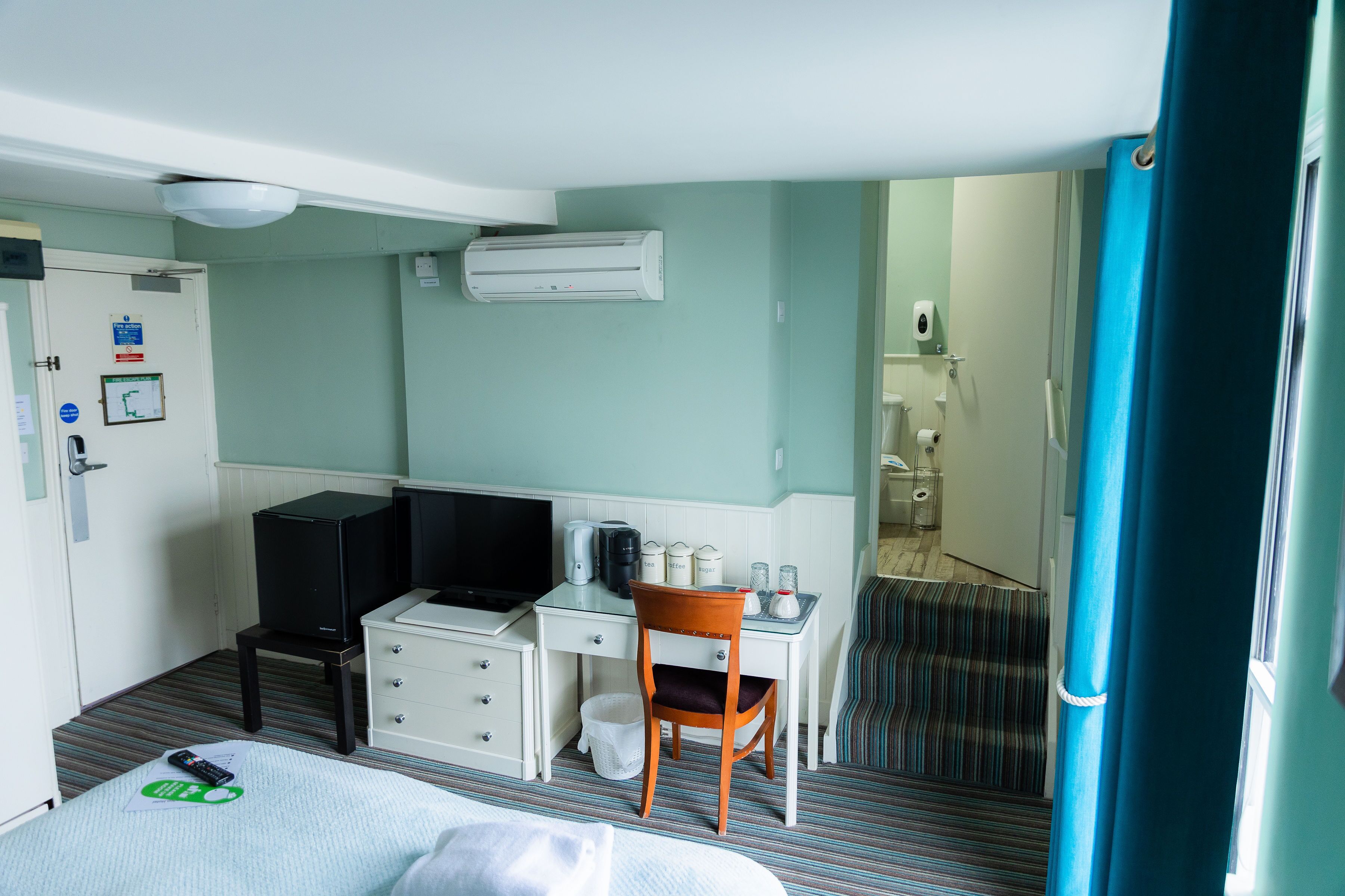 A hotel room with a bed, a desk with a chair, a television, a small table with a kettle, a trash bin, and a window with curtains.