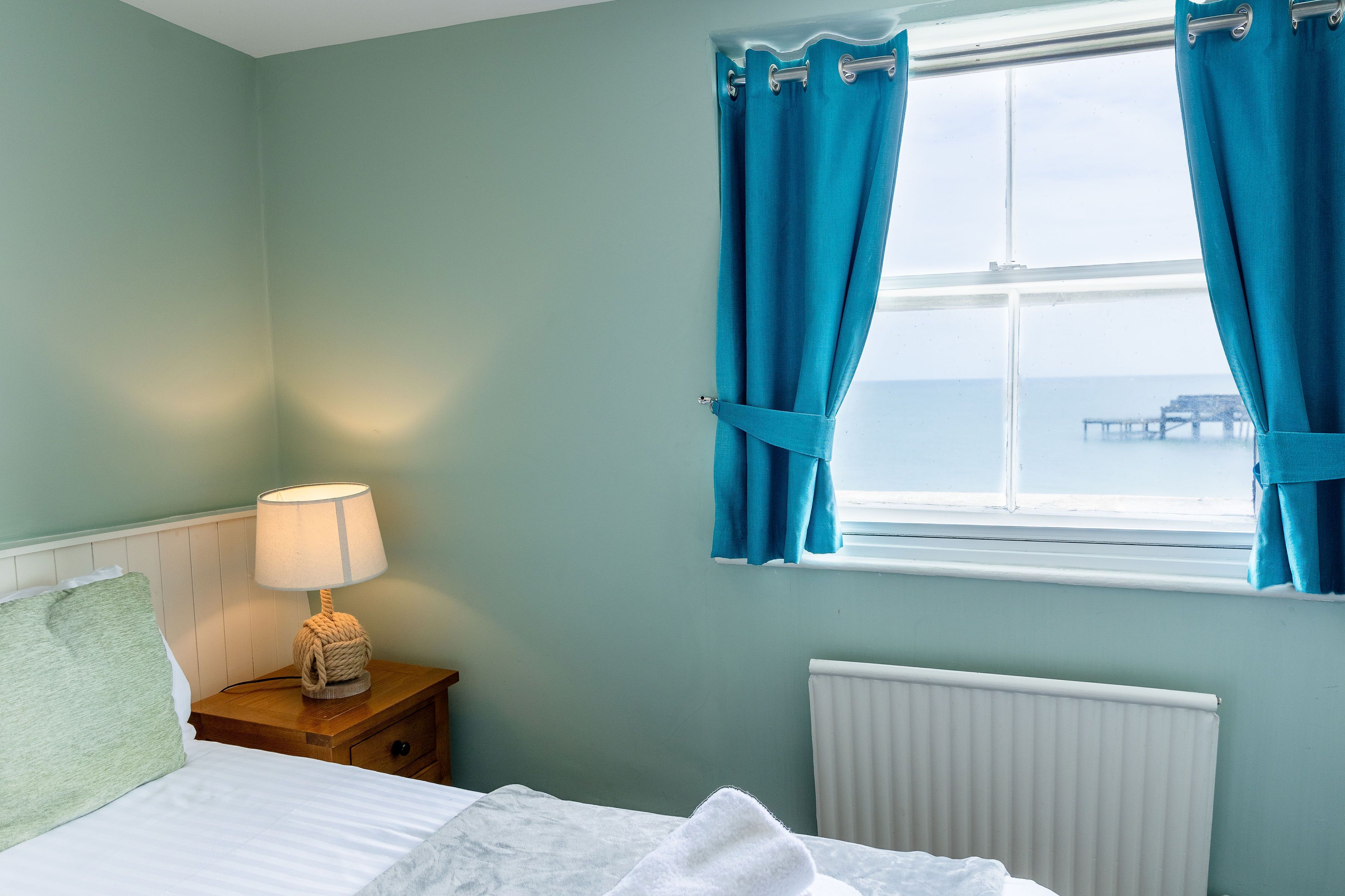 A bedroom with a bed, a nightstand with a lamp, a window with curtains, and a view of the sea.