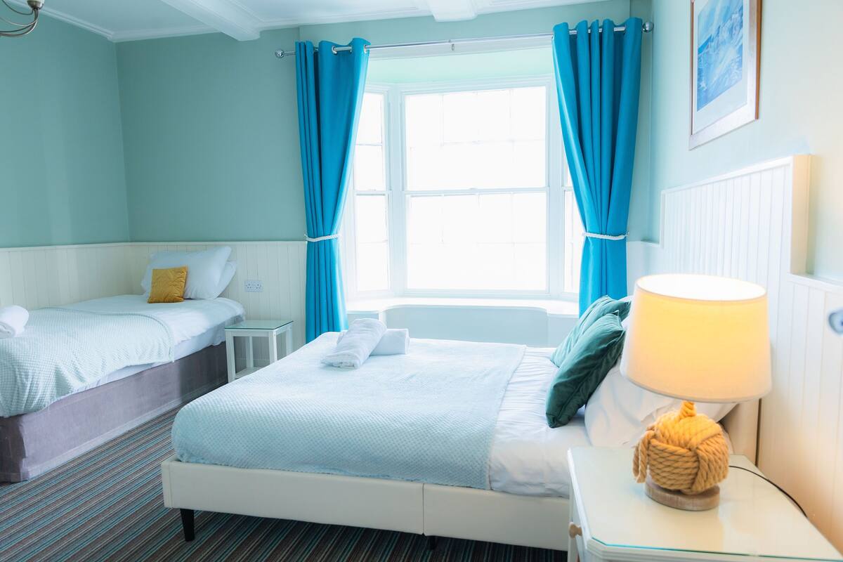 A hotel room with two beds, a window with curtains, a bedside lamp, and a framed picture on the wall.
