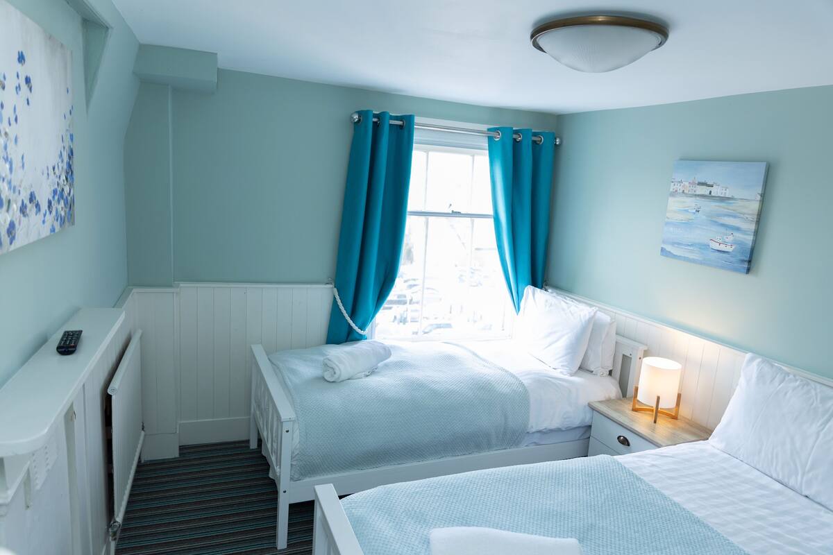 A twin bedroom with two beds, a nightstand with a lamp, a window with curtains, and a painting on the wall.