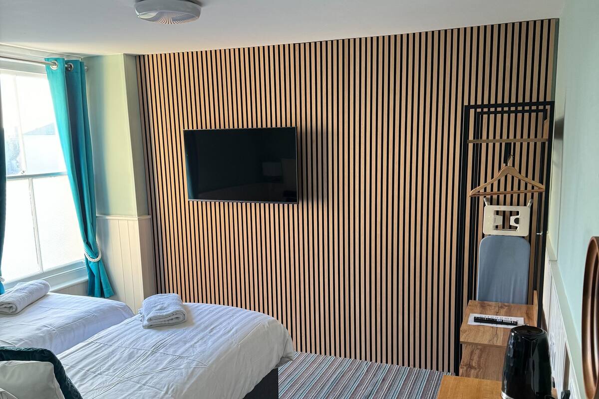 A hotel room with two beds, a TV mounted on the wall, a wooden side table, and a kettle.