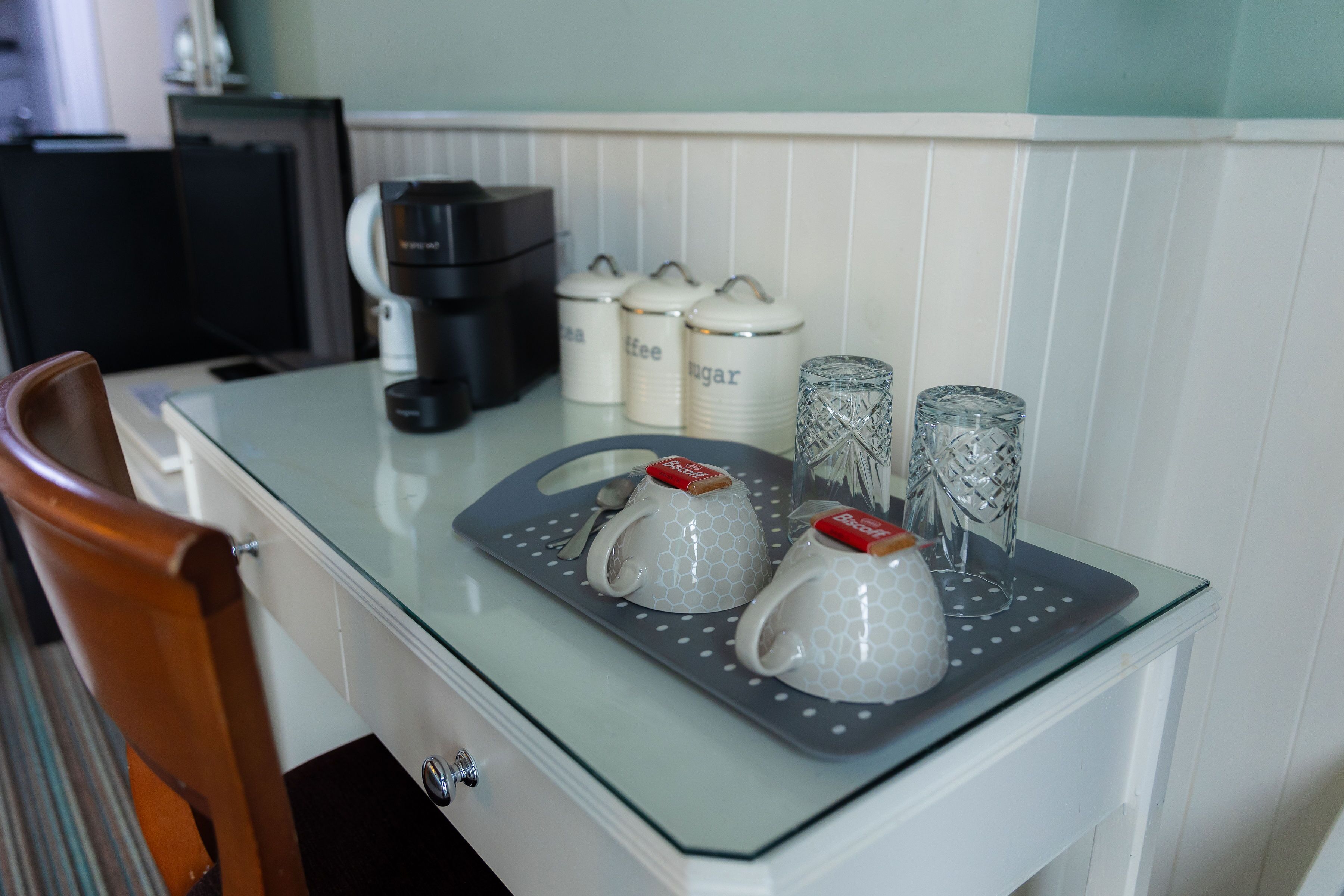 A kitchen counter with a coffee maker, sugar and cream containers, and two glass mugs with lids.