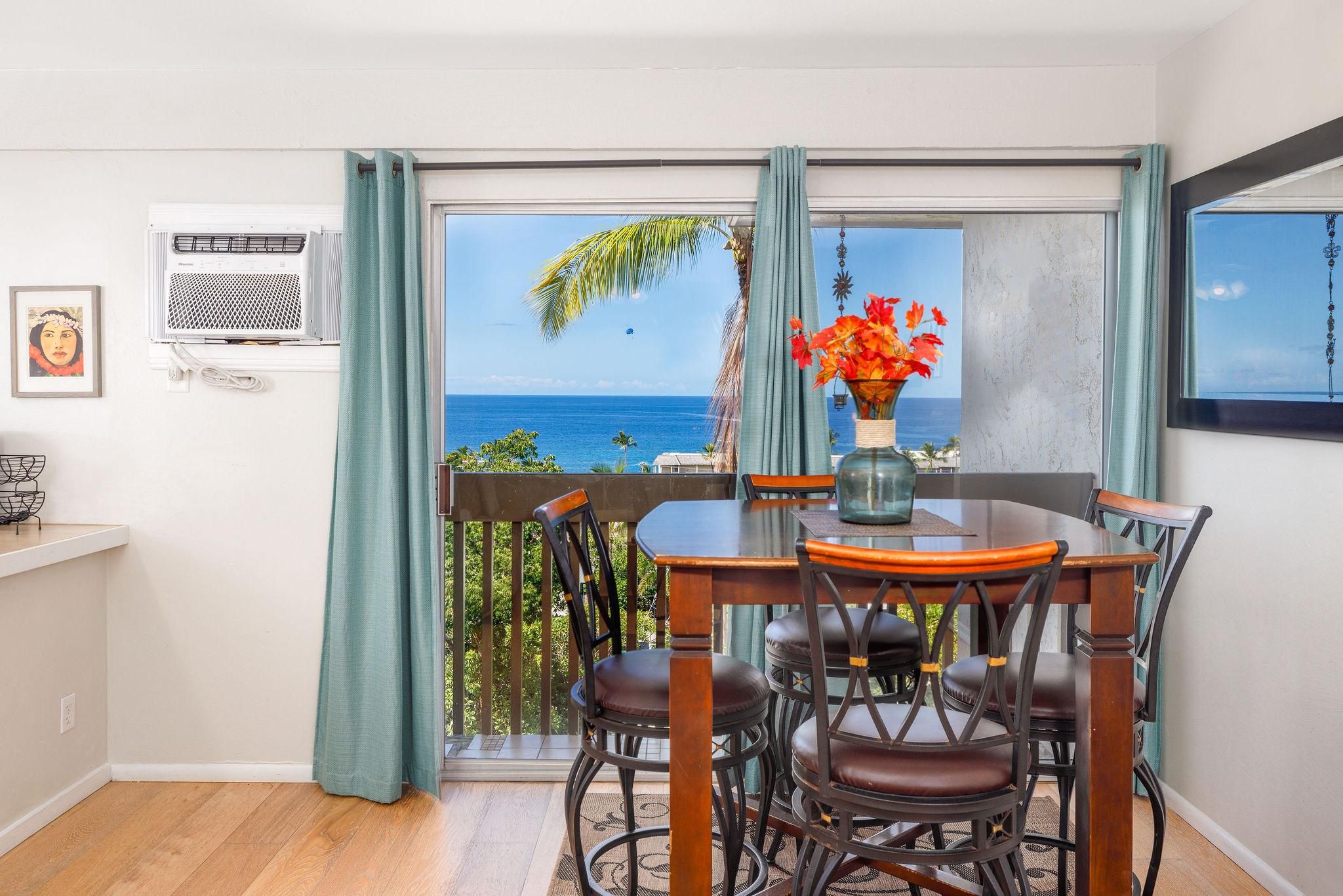 A dining area with a view of the ocean, a table with chairs, a vase with flowers, and a framed picture on the wall.