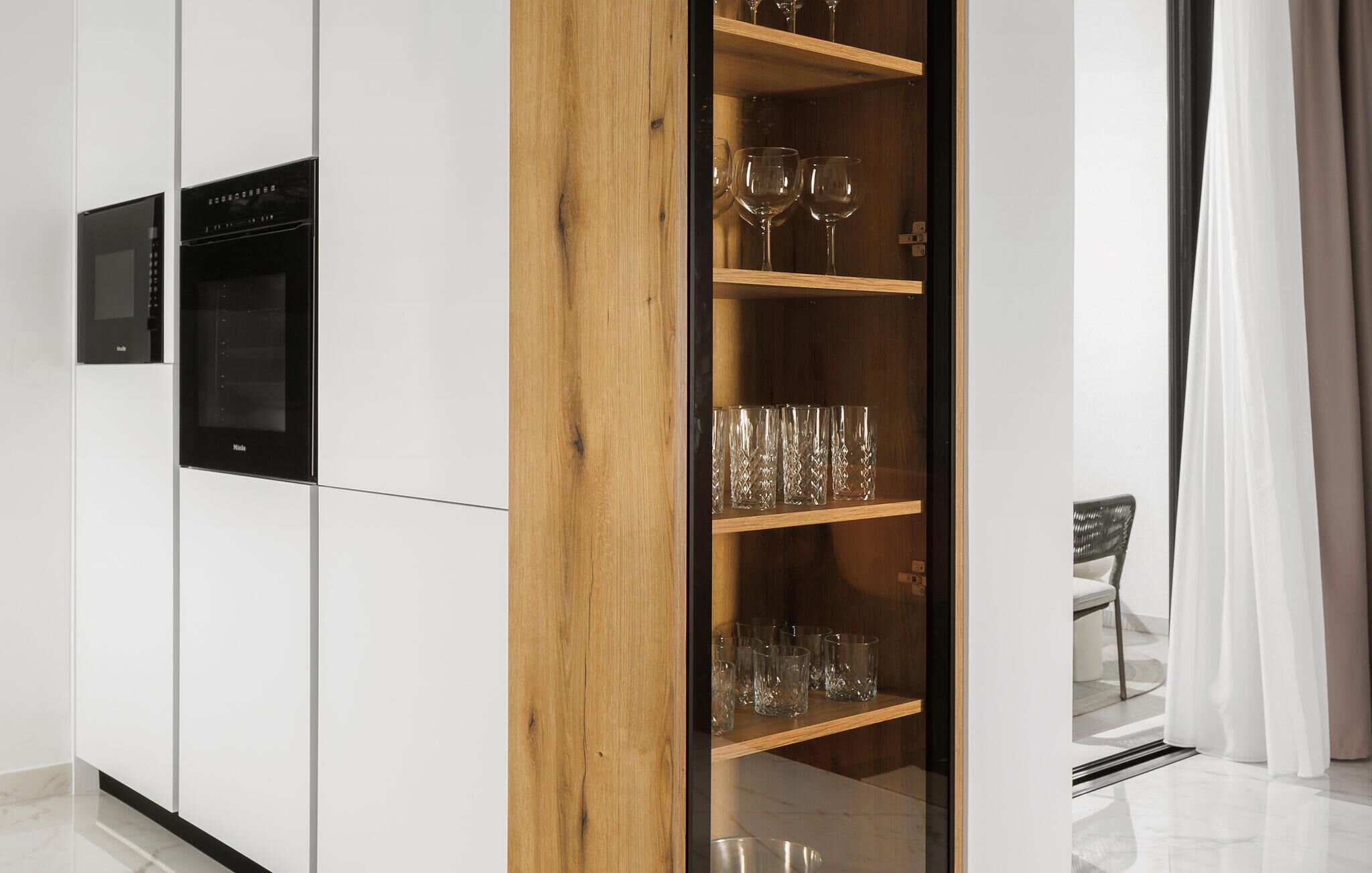 A modern kitchen with a built-in oven and microwave, wooden shelves with wine glasses, and a glass-fronted cabinet.