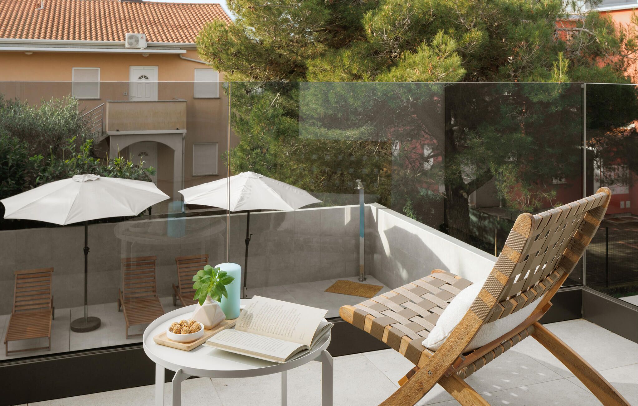 Ein Balkon mit Glastrennwand, einem Holzstuhl, einem Tisch mit einem Buch und einer Schüssel mit Snacks sowie zwei weiße Sonnenschirme.