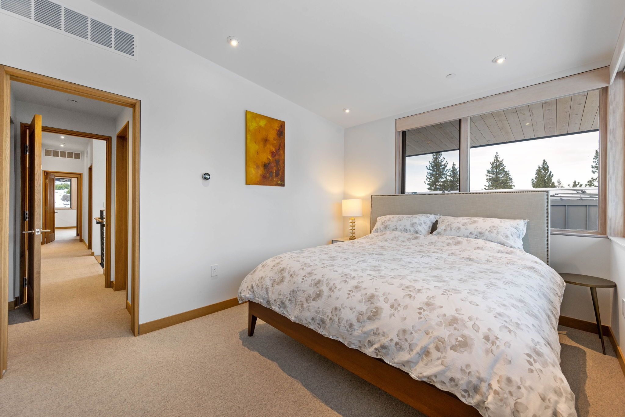 Une chambre avec un grand lit, une fenêtre donnant sur des arbres, un tableau accroché au mur et un couloir menant à une autre pièce.
