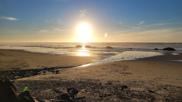 Ein Strand mit untergehendem Sonnenuntergang am Horizont, ruhigen Wellen und vereinzelten Felsen.
