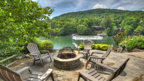 Une terrasse dotée de fauteuils Adirondack et d’un foyer extérieur, offrant une vue sur un lac où sont amarrés des bateaux, avec une cabane en arrière-plan.