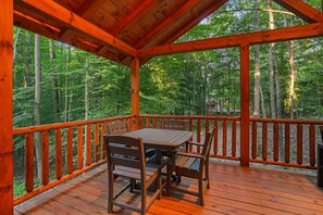 A wooden deck with a table and chairs, surrounded by a railing, overlooking a forested area.