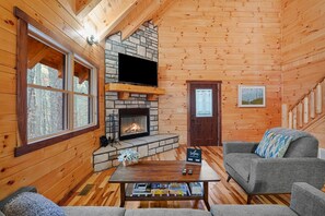 A cozy cabin interior with a stone fireplace, a television mounted above it, and a comfortable seating area with a sofa and armchairs.