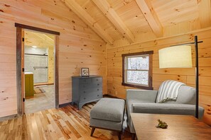 A cozy cabin interior with a wooden floor, a sofa, a small table, a dresser, and a window with a view of trees.