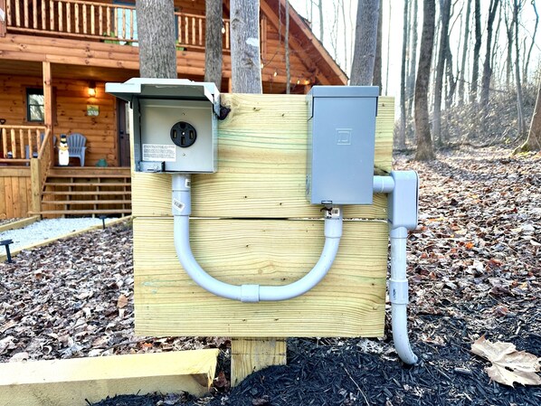 A wooden utility box with electrical components and a cable connected to it, situated outdoors with a wooden building in the background.