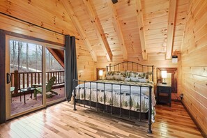 A wooden cabin bedroom with a bed, a nightstand, and a sliding glass door leading to an outdoor deck. - Green Frog | Luxury Cabin in the Woods, Hot Tub (Logan)