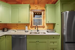 A kitchen with green cabinets, a stainless steel refrigerator, and a window with a view of trees. - Green Frog | Luxury Cabin in the Woods, Hot Tub (Logan)