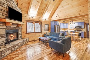 A cozy cabin interior with a stone fireplace, a sofa, a dining table, and a kitchen area. - Blue Bird | Luxury Cabin in the Woods (Logan)