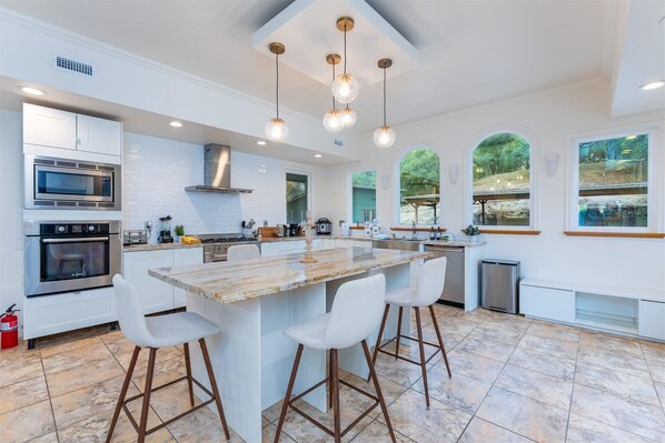 A modern kitchen with a central island, bar stools, stainless steel appliances, and large windows offering views of the outdoors.
