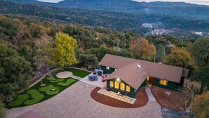 Une maison avec une allée pavée, une pelouse agrémentée d’un green d’entraînement au golf et une vue sur une chaîne de montagnes boisées.