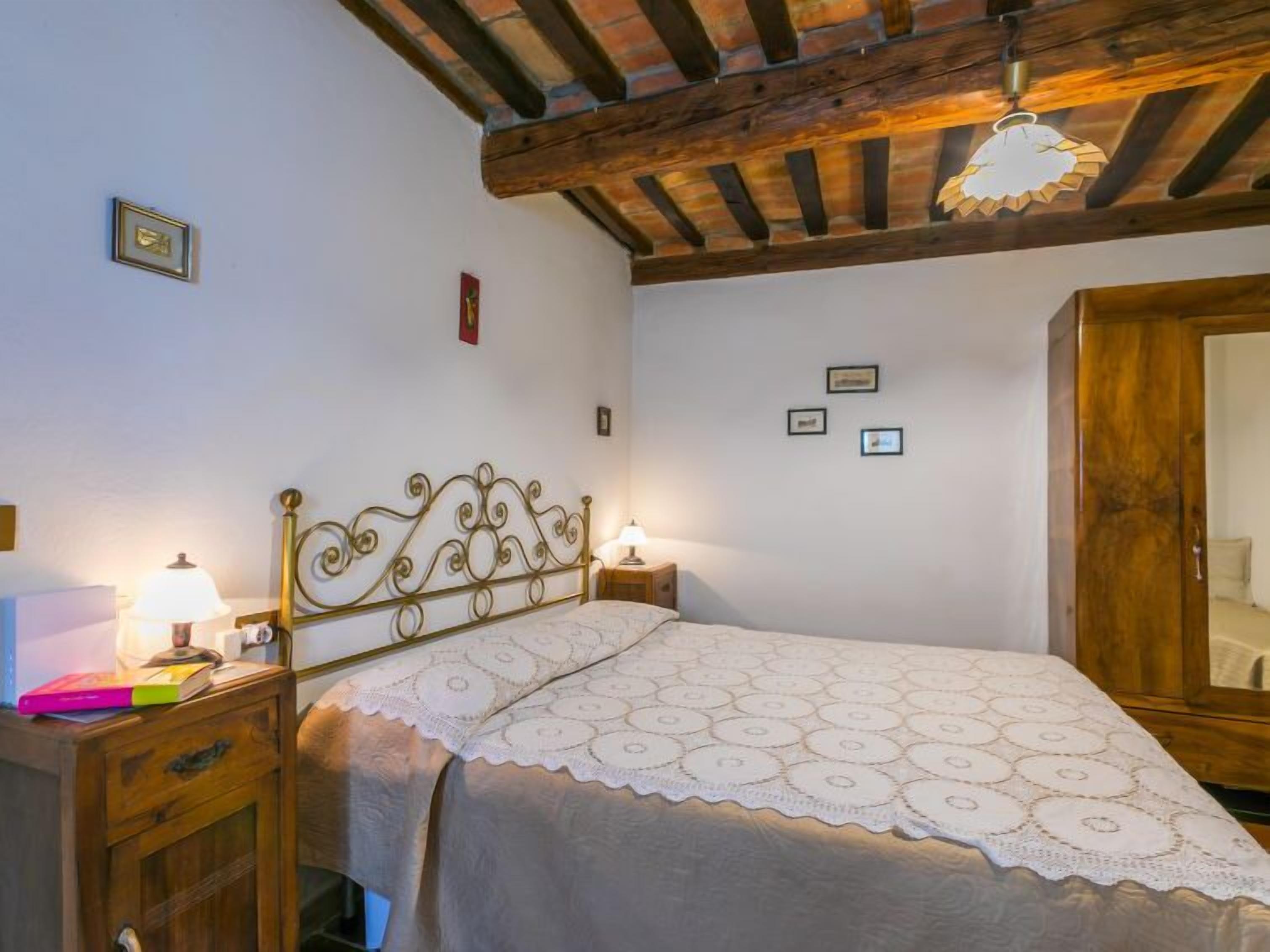 A bedroom with a bed, wooden headboard, bedside tables with lamps, a wardrobe, and a ceiling with exposed beams.