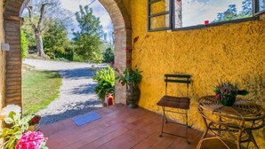 A covered patio with a brick floor, a wicker chair, a small table with a vase, and a view through an archway to a road and greenery. - Focolare by Interhome (Trequanda)